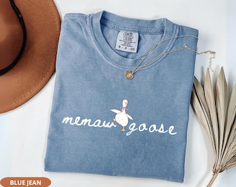 Pode incluir: Camiseta azul jeans Comfort Colors com o texto "memaw goose" e um desenho de ganso. Um colar de ouro com um pingente e um chapéu marrom estão ao lado da camiseta. Uma folha de palmeira seca também está na imagem.