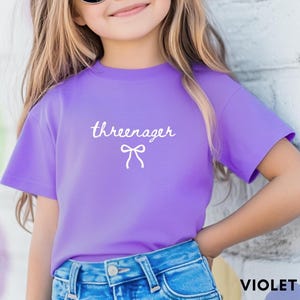May include: A violet t-shirt with the word "threenager" in cursive script and a bow graphic. The shirt is paired with blue denim jeans. The text "VIOLET COLOR" is at the bottom right.