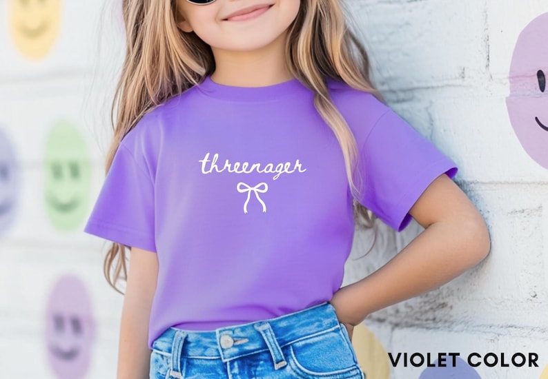 May include: A violet t-shirt with the word "threenager" in cursive script and a bow graphic. The shirt is paired with blue denim jeans. The text "VIOLET COLOR" is at the bottom right.