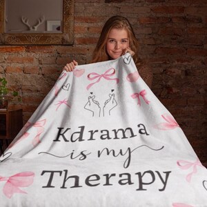 May include: White blanket with pink bows, hearts, and the text "Kdrama is my Therapy." The blanket features a hand gesture design. The blanket is held by a person.