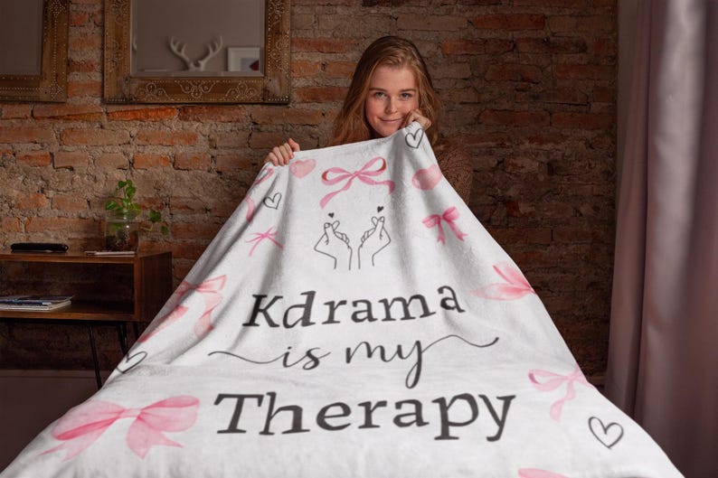 May include: White blanket with pink bows, hearts, and the text "Kdrama is my Therapy." The blanket features a hand gesture design. The blanket is held by a person.