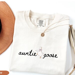 Puede incluir: Camiseta de color marfil con las palabras "auntie goose" en fuente cursiva, acompa&ntilde;ada de una ilustraci&oacute;n de un ganso de dibujos animados. Un collar dorado con un colgante descansa sobre la camiseta. La camiseta est&aacute; doblada sobre una superficie blanca.