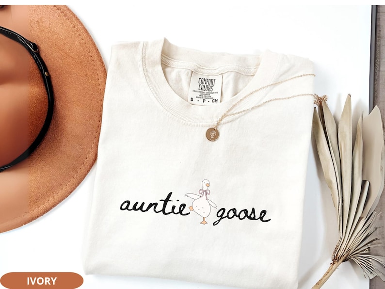 Puede incluir: Camiseta de color marfil con las palabras "auntie goose" en fuente cursiva, acompa&ntilde;ada de una ilustraci&oacute;n de un ganso de dibujos animados. Un collar dorado con un colgante descansa sobre la camiseta. La camiseta est&aacute; doblada sobre una superficie blanca.