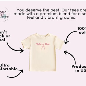 May include: Cream-colored t-shirt with the text "Child of God" in pink script. The shirt is made of 100% US cotton and is produced in the USA. The graphic is designed to be ultra-comfortable and won't crack or peel.