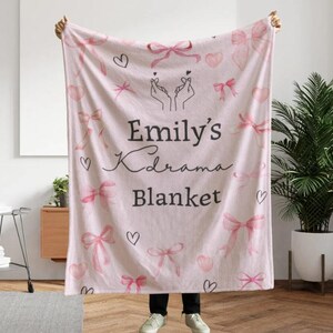May include: A light pink blanket with a pattern of pink bows, hearts, and the text "Emily's Kdrama Blanket." The blanket is held up by a person. The blanket has a soft, cozy appearance.