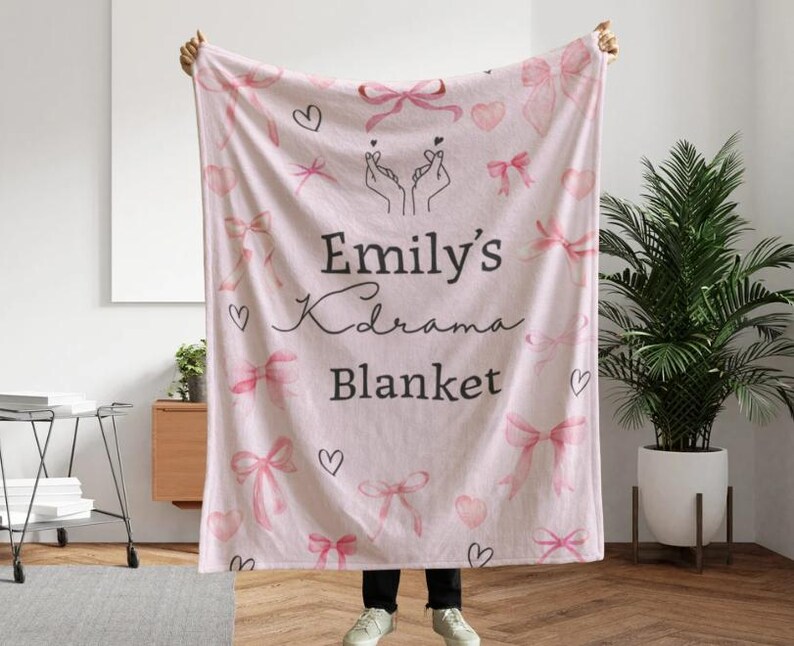May include: A light pink blanket with a pattern of pink bows, hearts, and the text "Emily's Kdrama Blanket." The blanket is held up by a person. The blanket has a soft, cozy appearance.