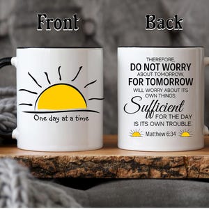 May include: White ceramic mug with a black handle and rim. The front of the mug has a black and yellow sun graphic with the text "One day at a time". The back of the mug has black text that reads "Therefore, do not worry about tomorrow, for tomorrow will worry about its own things. Sufficient for the day is its own trouble. - Matthew 6:34".