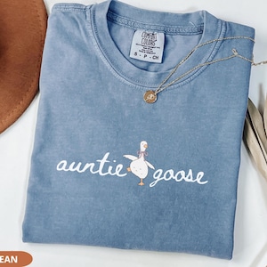 Puede incluir: Una camiseta azul vaquera Comfort Colors con las palabras "auntie goose" y un dibujo de un ganso. Un collar dorado con un colgante est&aacute; sobre la camiseta. Un sombrero marr&oacute;n y una planta seca est&aacute;n en el fondo.