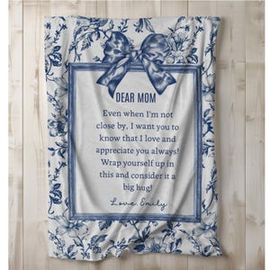 Pode incluir: Um cobertor branco com um padr&atilde;o floral azul e um design de la&ccedil;o azul. O cobertor tem uma mensagem emoldurada que diz "DEAR MOM" e uma mensagem pessoal. O cobertor &eacute; um presente atencioso.
