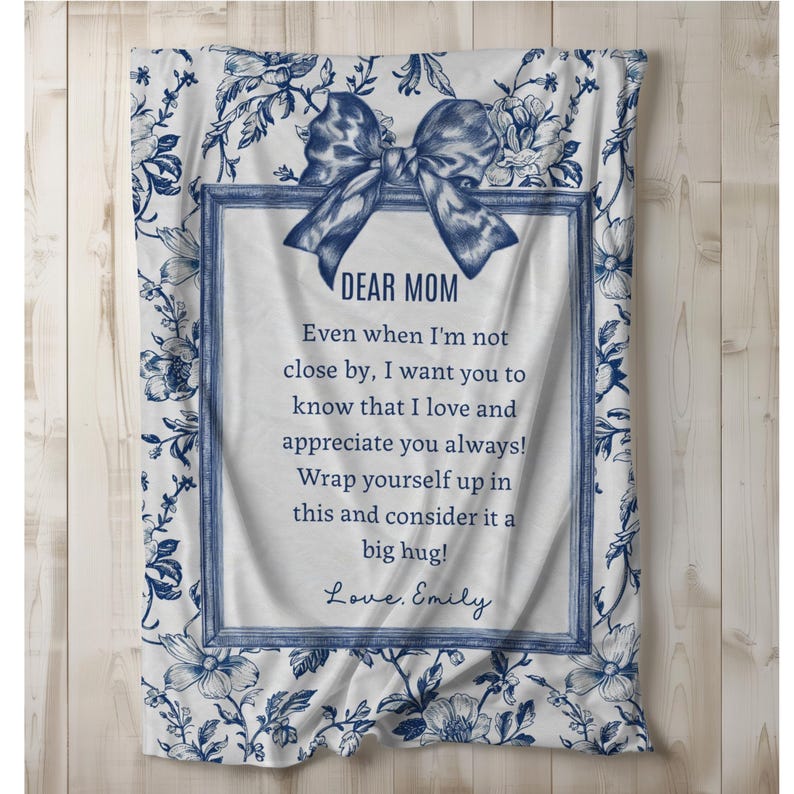 Pode incluir: Um cobertor branco com um padr&atilde;o floral azul e um design de la&ccedil;o azul. O cobertor tem uma mensagem emoldurada que diz "DEAR MOM" e uma mensagem pessoal. O cobertor &eacute; um presente atencioso.