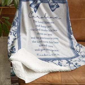 May include: A white and blue floral throw blanket draped over a brown leather chair. The blanket features a bow design and the name "Caroline" in script, along with a Bible verse. The blanket has a soft, plush texture.
