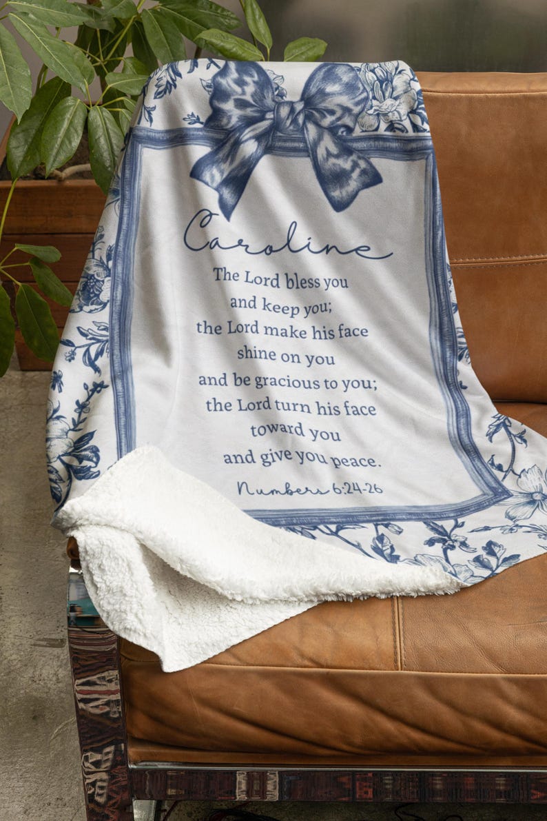May include: A white and blue floral throw blanket draped over a brown leather chair. The blanket features a bow design and the name "Caroline" in script, along with a Bible verse. The blanket has a soft, plush texture.
