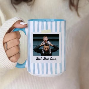 May include: White ceramic mug with a blue interior and handle. The mug features a light blue vertical stripe pattern and a photo with the text "Best Dad Ever". The photo shows a man with a child on his shoulders.