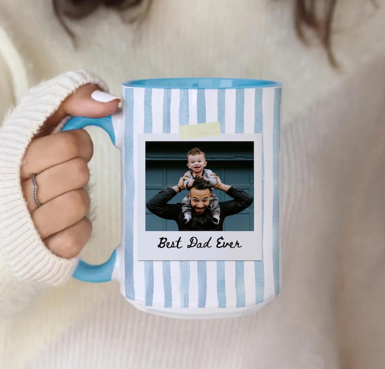 May include: White ceramic mug with a blue interior and handle. The mug features a light blue vertical stripe pattern and a photo with the text "Best Dad Ever". The photo shows a man with a child on his shoulders.
