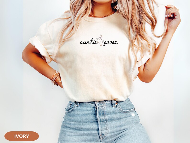 Puede incluir: Camiseta de color marfil con la frase "auntie goose" en escritura negra. El dise&ntilde;o incluye una sencilla ilustraci&oacute;n de un ganso. La camiseta se combina con vaqueros azules.