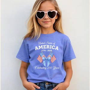 May include: A Columbia blue t-shirt with the text "United States of AMERICA 1776-2026 Celebrating 250 Years" and two American flags. The shirt is worn by a person wearing heart-shaped sunglasses.