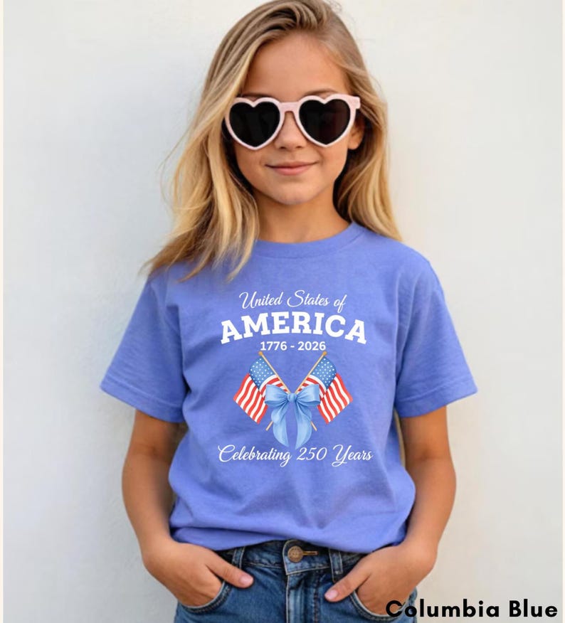 May include: A Columbia blue t-shirt with the text "United States of AMERICA 1776-2026 Celebrating 250 Years" and two American flags. The shirt is worn by a person wearing heart-shaped sunglasses.