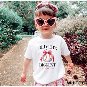 Puede incluir: Una ni&ntilde;a lleva una camiseta blanca con un dise&ntilde;o de b&eacute;isbol y el texto "Oliver's Biggest Fan". La camiseta presenta una pelota de b&eacute;isbol con un lazo rojo. La ni&ntilde;a lleva gafas de sol rosas en forma de coraz&oacute;n y un lazo rosa en el pelo.