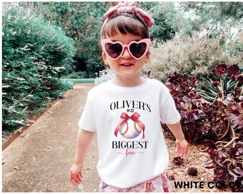 Puede incluir: Una ni&ntilde;a lleva una camiseta blanca con un dise&ntilde;o de b&eacute;isbol y el texto "Oliver's Biggest Fan". La camiseta presenta una pelota de b&eacute;isbol con un lazo rojo. La ni&ntilde;a lleva gafas de sol rosas en forma de coraz&oacute;n y un lazo rosa en el pelo.