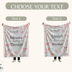 May include: A white blanket with pink bows, hearts, and the text "Kdrama is my Therapy". The blanket also features a hand gesture and is held up by a person. The blanket is a soft, cozy item.