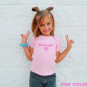 May include: A pink t-shirt with the word "threenager" in a cursive font and a bow graphic. The shirt is worn by a young girl, paired with blue jeans and a blue scrunchie. The background is a white textured wall.