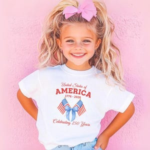 May include: A white t-shirt with the text "United States of AMERICA" in red, along with the years 1776-2026, and "Celebrating 250 Years" in red. The shirt features two American flags and a blue bow. A young girl is wearing the shirt.