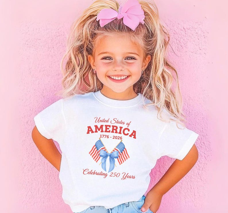 May include: A white t-shirt with the text "United States of AMERICA" in red, along with the years 1776-2026, and "Celebrating 250 Years" in red. The shirt features two American flags and a blue bow. A young girl is wearing the shirt.