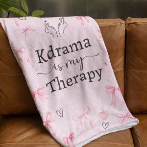 May include: A light pink blanket with the text "Kdrama is my Therapy" in black script. The blanket features pink bows and hearts. The blanket is folded over a brown leather couch.