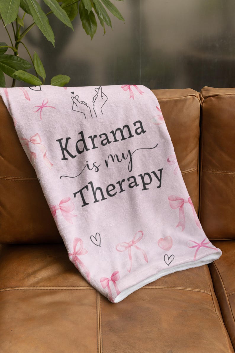 May include: A light pink blanket with the text "Kdrama is my Therapy" in black script. The blanket features pink bows and hearts. The blanket is folded over a brown leather couch.