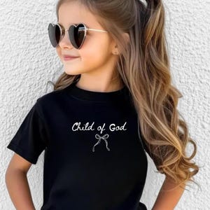 May include: Black t-shirt with the text "Child of God" in white script and a bow graphic. The shirt is short-sleeved and has a round neckline. The image also shows heart-shaped sunglasses.
