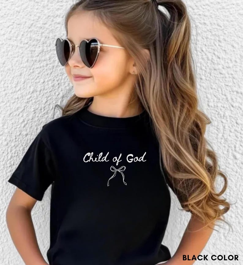 May include: Black t-shirt with the text "Child of God" in white script and a bow graphic. The shirt is short-sleeved and has a round neckline. The image also shows heart-shaped sunglasses.