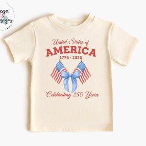 May include: Cream-colored t-shirt featuring the text "United States of AMERICA" in red, with the years 1776-2026. Two American flags and a blue bow are below the text, with the words "Celebrating 250 Years" in red.
