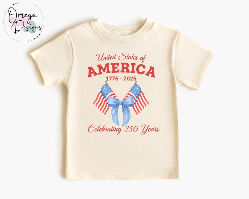 May include: Cream-colored t-shirt featuring the text "United States of AMERICA" in red, with the years 1776-2026. Two American flags and a blue bow are below the text, with the words "Celebrating 250 Years" in red.