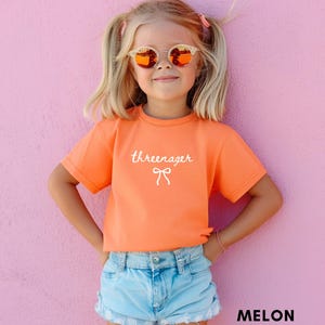 May include: A young girl wearing an orange t-shirt with the word "threeenager" and a bow graphic. She is also wearing light blue denim shorts and sunglasses. The background is a solid pink color.