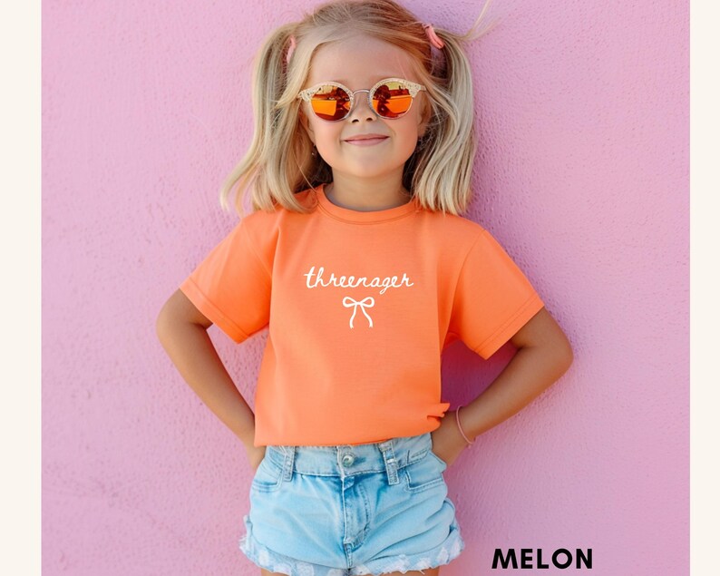 May include: A young girl wearing an orange t-shirt with the word "threeenager" and a bow graphic. She is also wearing light blue denim shorts and sunglasses. The background is a solid pink color.