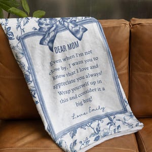 Pode incluir: Um cobertor branco com um design floral azul e o texto "DEAR MOM" em uma moldura decorativa. O cobertor inclui uma mensagem e est&aacute; dobrado em um sof&aacute; de couro marrom.
