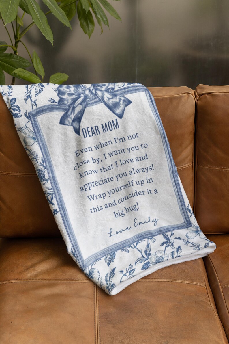 Pode incluir: Um cobertor branco com um design floral azul e o texto "DEAR MOM" em uma moldura decorativa. O cobertor inclui uma mensagem e est&aacute; dobrado em um sof&aacute; de couro marrom.