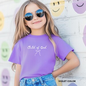 May include: A violet-colored t-shirt with the text "Child of God" and a bow graphic. The shirt is worn by a young person wearing sunglasses and blue jeans. The words "VIOLET COLOR" are at the bottom right.