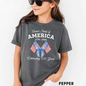 May include: A dark gray t-shirt featuring the text "United States of AMERICA 1776-2026 Celebrating 250 Years." The design includes two American flags tied with a blue bow. The shirt is a comfortable, casual style.