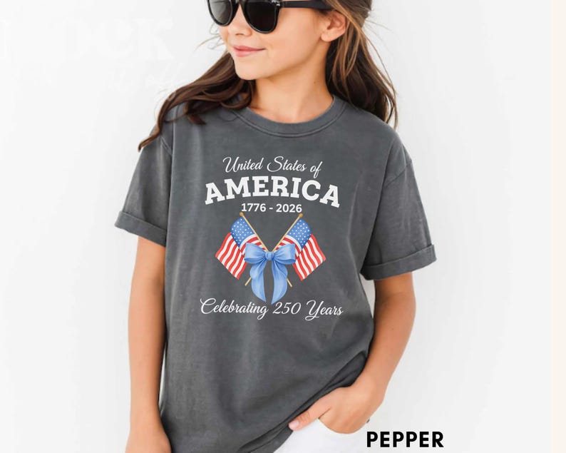 May include: A dark gray t-shirt featuring the text "United States of AMERICA 1776-2026 Celebrating 250 Years." The design includes two American flags tied with a blue bow. The shirt is a comfortable, casual style.