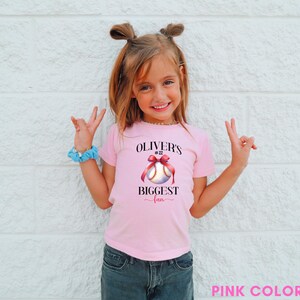 Puede incluir: Una camiseta rosa con el texto "OLIVER'S BIGGEST fan" y un gr&aacute;fico de b&eacute;isbol con un lazo rojo. La camiseta la lleva una ni&ntilde;a con el pelo recogido en dos mo&ntilde;os, con vaqueros azules y una goma de pelo azul.