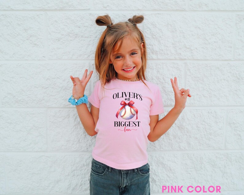 Puede incluir: Una camiseta rosa con el texto "OLIVER'S BIGGEST fan" y un gr&aacute;fico de b&eacute;isbol con un lazo rojo. La camiseta la lleva una ni&ntilde;a con el pelo recogido en dos mo&ntilde;os, con vaqueros azules y una goma de pelo azul.
