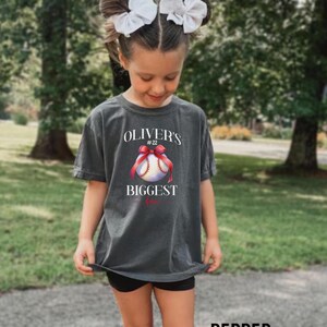 Puede incluir: Una camiseta gris carb&oacute;n con el texto "Oliver's Biggest Fan" y un gr&aacute;fico de b&eacute;isbol con un lazo rojo. La camiseta la lleva una persona joven con lazos blancos en el pelo.