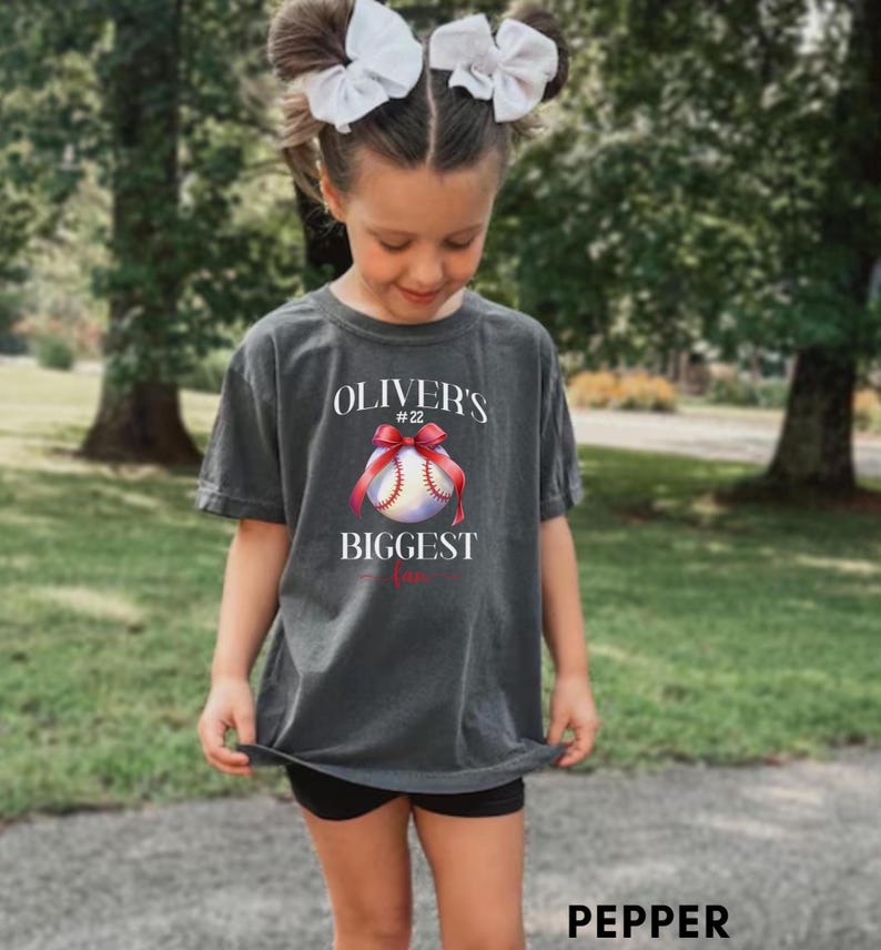 Puede incluir: Una camiseta gris carb&oacute;n con el texto "Oliver's Biggest Fan" y un gr&aacute;fico de b&eacute;isbol con un lazo rojo. La camiseta la lleva una persona joven con lazos blancos en el pelo.
