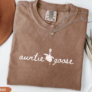 Puede incluir: Camiseta marr&oacute;n con las palabras "auntie goose" y un dibujo de un ganso. La camiseta tiene cuello redondo y mangas cortas. Un collar plateado con un colgante est&aacute; sobre la camiseta. Un sombrero marr&oacute;n es visible en el fondo.