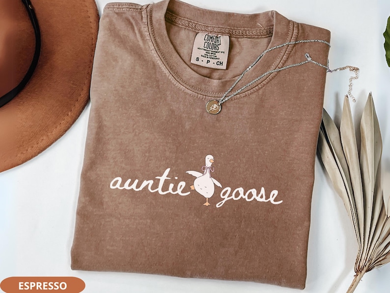 Puede incluir: Camiseta marr&oacute;n con las palabras "auntie goose" y un dibujo de un ganso. La camiseta tiene cuello redondo y mangas cortas. Un collar plateado con un colgante est&aacute; sobre la camiseta. Un sombrero marr&oacute;n es visible en el fondo.