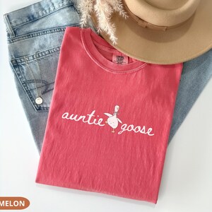 Puede incluir: Una camiseta de color sand&iacute;a con las palabras "auntie goose" y un gr&aacute;fico de ganso. La camiseta est&aacute; colocada sobre un par de vaqueros azul claro y un sombrero fedora marr&oacute;n. Se ve un collar dorado.