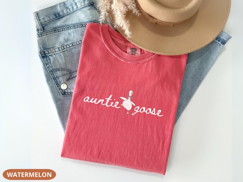 Puede incluir: Una camiseta de color sand&iacute;a con las palabras "auntie goose" y un gr&aacute;fico de ganso. La camiseta est&aacute; colocada sobre un par de vaqueros azul claro y un sombrero fedora marr&oacute;n. Se ve un collar dorado.