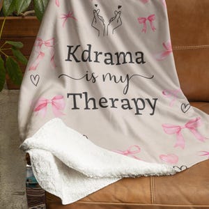May include: A beige blanket with a white sherpa lining, featuring pink bows, hearts, and the text "Kdrama is my Therapy" in black script. The blanket is draped over a brown leather couch.