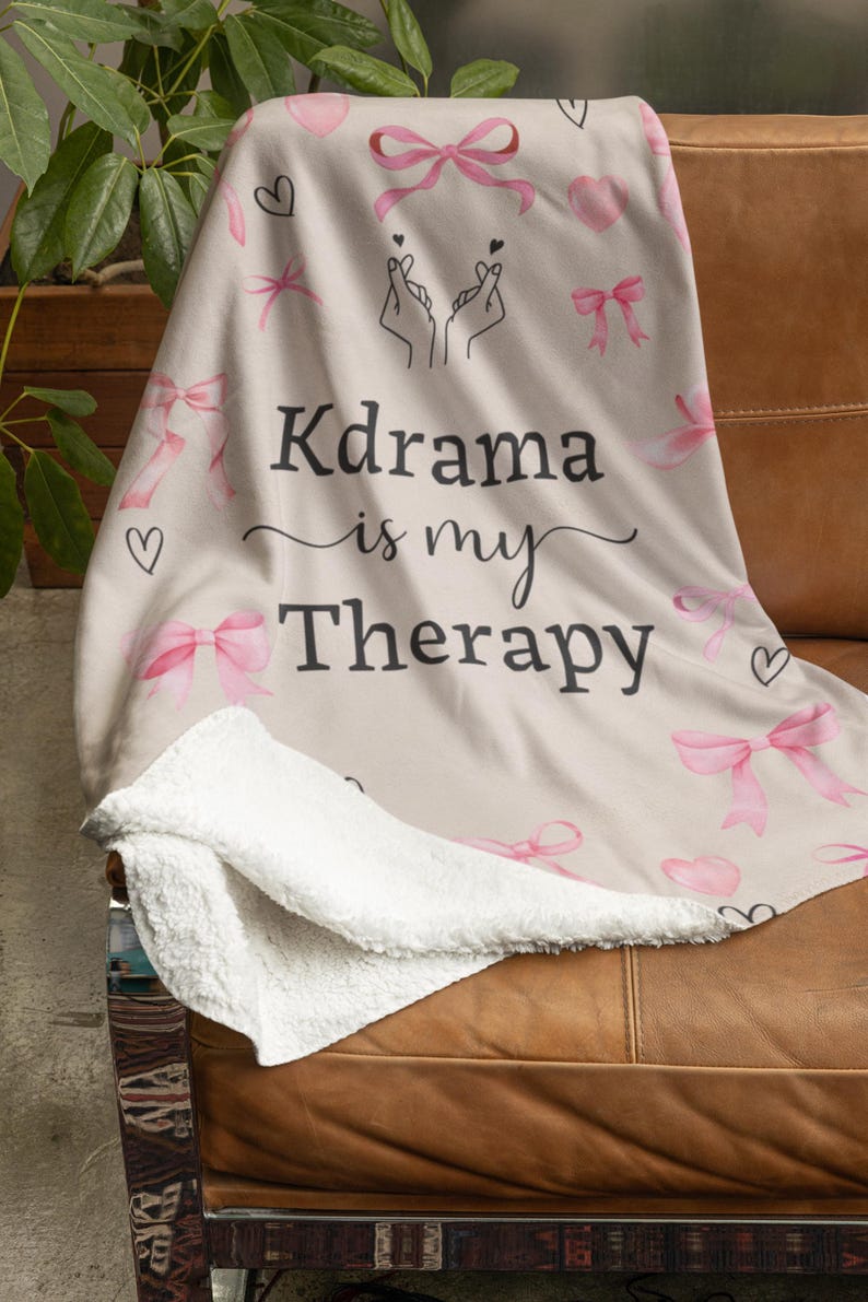 May include: A beige blanket with a white sherpa lining, featuring pink bows, hearts, and the text "Kdrama is my Therapy" in black script. The blanket is draped over a brown leather couch.
